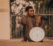 A contemplative man in a suit sits outdoors holding a large clock, embodying the passage of time.