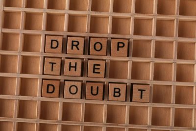 Wooden letter tiles spelling 'Drop the Doubt' on a wooden grid background, promoting positivity.