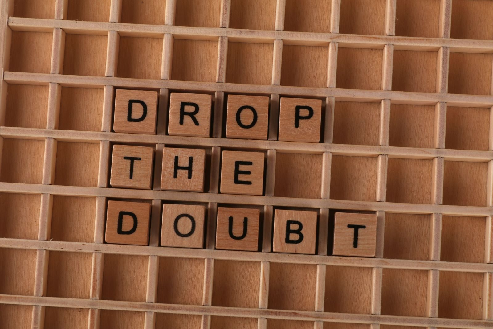 Wooden letter tiles spelling 'Drop the Doubt' on a wooden grid background, promoting positivity.